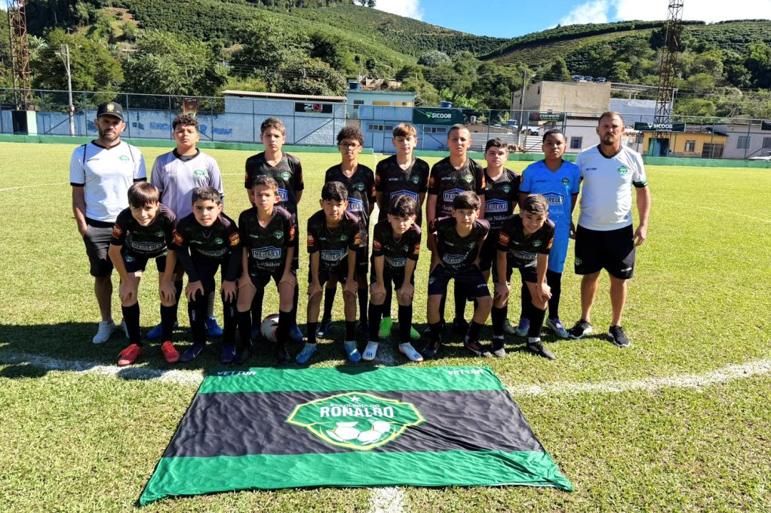 Foto oficial da equipe Sub-13 da Escolinha do Ronaldo reunida no gramado. À frente dos jogadores, estendida no chão, está uma grande bandeira nas cores preta e verde com o escudo do time escrito 