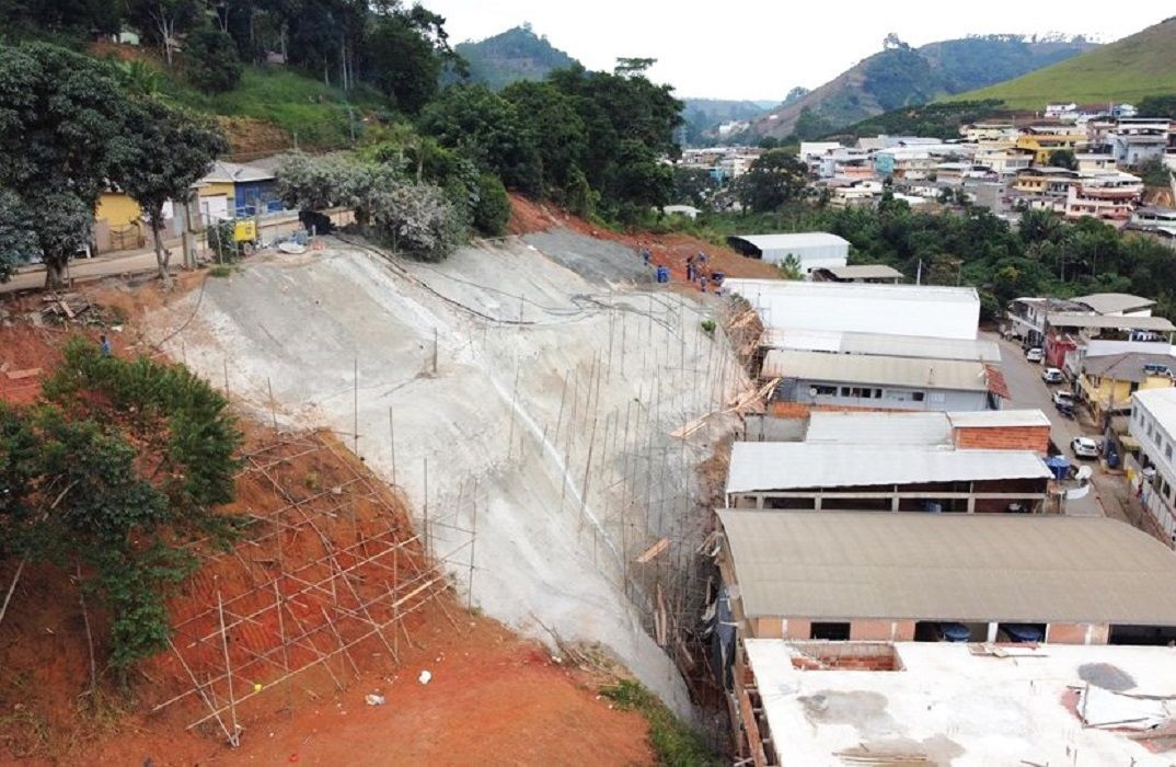 Vista aérea de encosta urbana em obras no município de Ibitirama. Um grande talude de terra está parcialmente coberto por concreto projetado (solo grampeado), com andaimes de madeira e estruturas de metal instalados ao longo da face da encosta. Na parte superior, há vegetação e uma rua residencial; na parte inferior e lateral direita, observam-se telhados de casas e galpões comerciais próximos à base da encosta. Ao fundo, aparecem montanhas características da região do Caparaó.