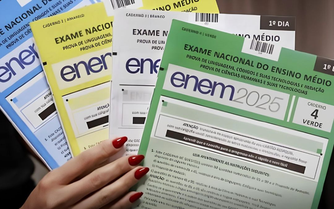 Vista aproximada das mãos de uma pessoa com unhas pintadas de vermelho segurando quatro cadernos de prova oficiais do Enem, dispostos em leque. Os cadernos apresentam capas em cores diferentes: azul, amarelo, branco e verde (este último em destaque na frente, indicando 'Caderno 4 - Verde'). Os livretos trazem o logotipo do exame impresso na parte superior.