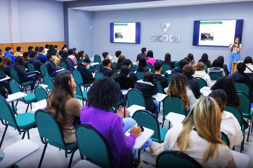 Vista geral do auditório do Sicoob em Iúna, lotado de estudantes sentados em cadeiras estofadas verdes com pranchetas. Ao fundo, uma palestrante de calça azul e blusa branca fala ao microfone ao lado de duas telas de TV que exibem slides com o título 
