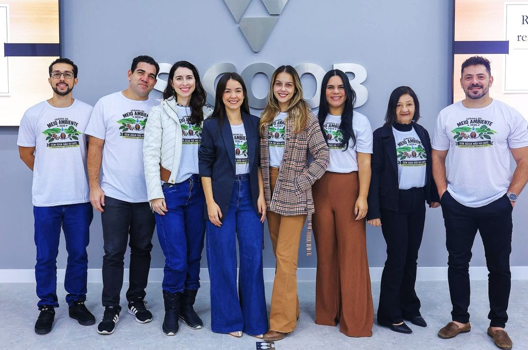 Foto oficial da equipe organizadora alinhada lado a lado em frente à parede cinza com a marca Sicoob. O grupo é composto por cinco mulheres e três homens. Quatro deles vestem a camiseta oficial branca do evento com a estampa 