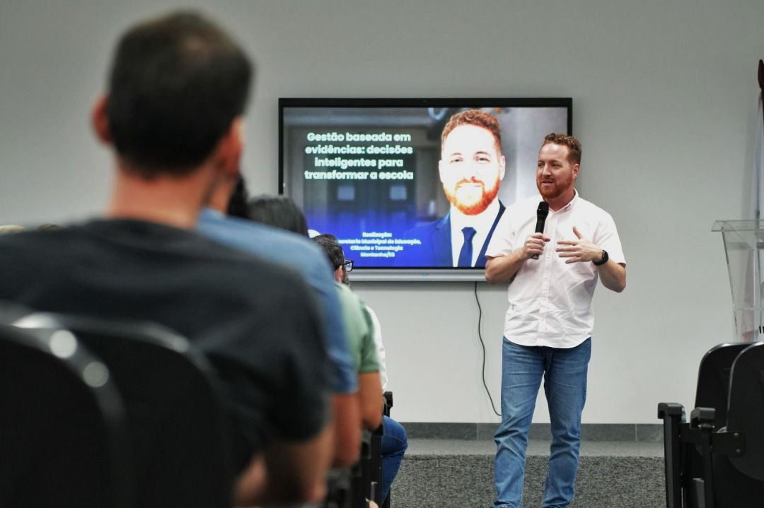 Fotografia em plano médio de Vitor de Angelo durante uma palestra. Ele é um homem de pele clara, cabelos ruivos curtos e barba ruiva aparada, veste camisa social branca de mangas curtas e calça jeans azul clara. Ele está de pé, no lado direito da imagem, segurando um microfone preto na mão direita e gesticulando com a mão esquerda enquanto fala. Ao fundo, fixada na parede cinza, há uma tela de TV digital exibindo um slide com sua própria foto oficial de terno e gravata e o texto: 