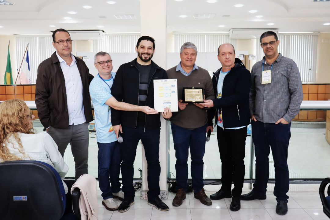 Sete homens adultos em pé, alinhados lado a lado no interior da Câmara Municipal de Iúna. Ao centro, dois homens seguram conjuntamente uma placa de homenagem escura com letras douradas e uma folha de papel impressa. Parte do grupo veste camisetas azuis com a estampa 