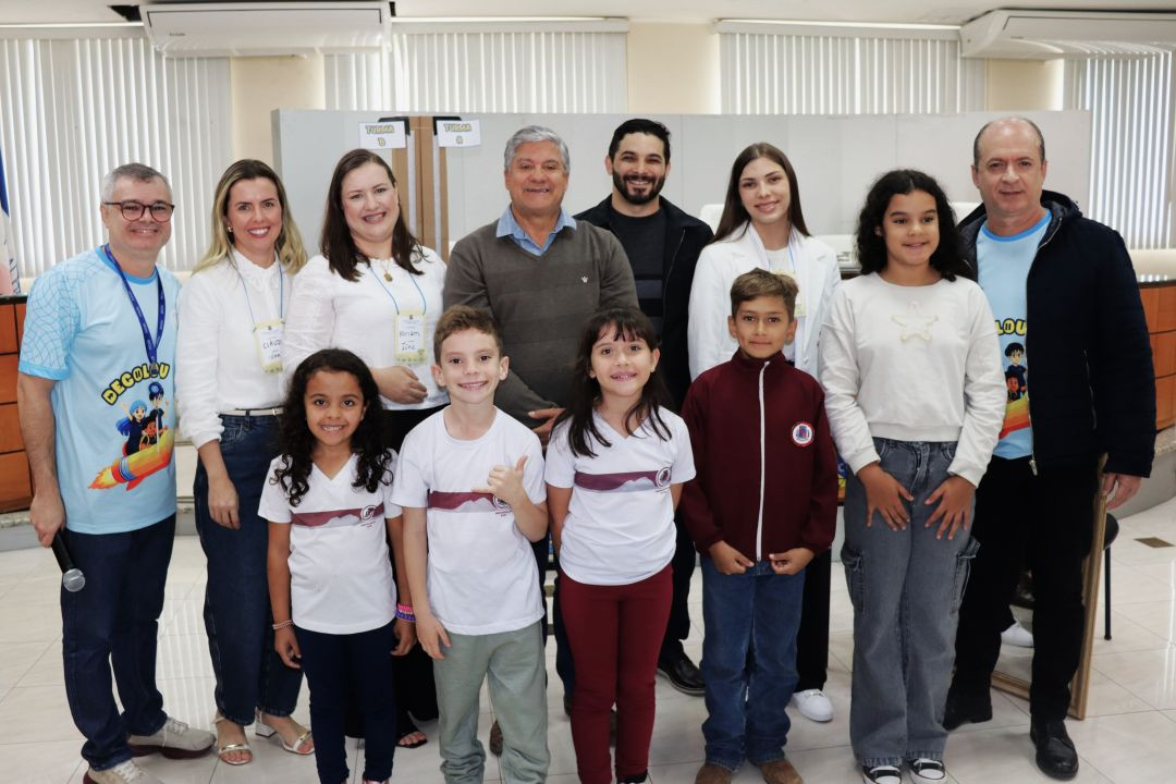 Foto em grupo com cinco adultos ao fundo e cinco crianças à frente, posicionados em ambiente interno iluminado. Os adultos sorriem; dois deles usam camisetas azuis com estampa escrita 