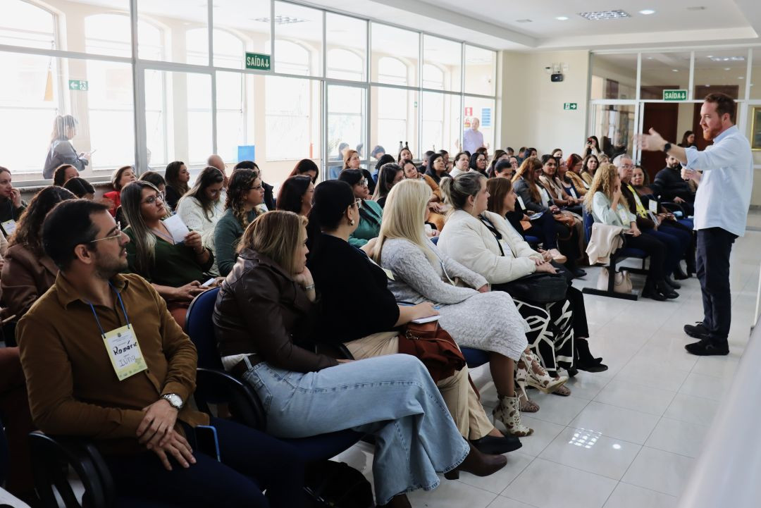 Visão lateral de um auditório repleto de pessoas sentadas em cadeiras azuis, a maioria mulheres, prestando atenção a uma palestra. No primeiro plano, à esquerda, um homem com crachá onde se lê 