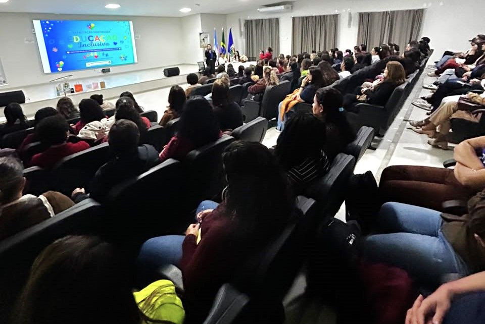 Visão panorâmica de cima das fileiras de cadeiras pretas do Auditório Francimar Carlos Pinheiro, ocupadas por dezenas de profissionais de educação de costas. Ao fundo, no palco iluminado, há um grande telão azul com o título 