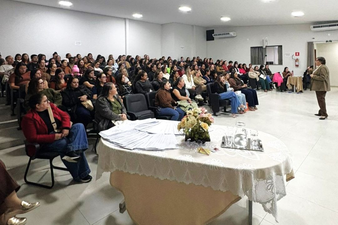 Vista lateral da plateia sentada nas cadeiras do auditório, preenchendo o espaço. Em primeiro plano, uma mesa coberta com toalha clara rendada exibe apostilas e papéis organizados, além de um vaso com flores e uma jarra de água. No canto direito, a palestrante Karla Coelho, vestindo blazer marrom e calça escura, gesticula enquanto ministra a formação voltada para o público.