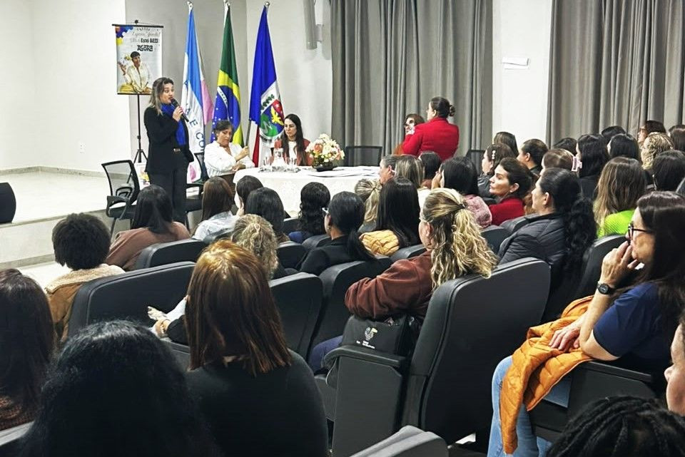 Plano médio focado na área do palco do auditório. À esquerda, uma mulher de blazer preto fala ao microfone em pé. Ao centro, atrás de uma mesa com flores e documentos, estão sentadas três mulheres, incluindo a secretária de Educação Esther Simões. Ao fundo, estão posicionadas três bandeiras oficiais em pedestais (do Espírito Santo, do Brasil e do município). A plateia aparece em primeiro plano, sentada nas poltronas escuras.