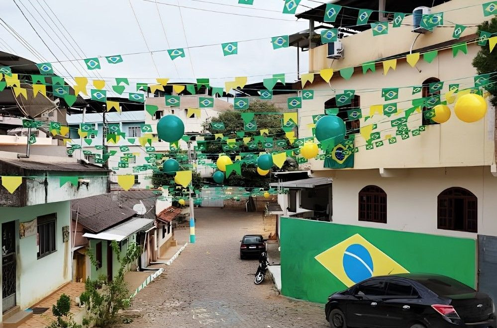 Vista longitudinal de uma rua residencial de calçamento de pedras no distrito de Piaçu. No alto, cruzando a via de um lado ao outro, estão penduradas fileiras de bandeirinhas verdes com o brasão do Brasil e balões infláveis redondos nas cores verde e amarela. À direita, um muro verde exibe a pintura estilizada da bandeira do Brasil, posicionado ao lado de um carro preto estacionado. Ao fundo, veem-se casas e sobrados sob um céu nublado.
