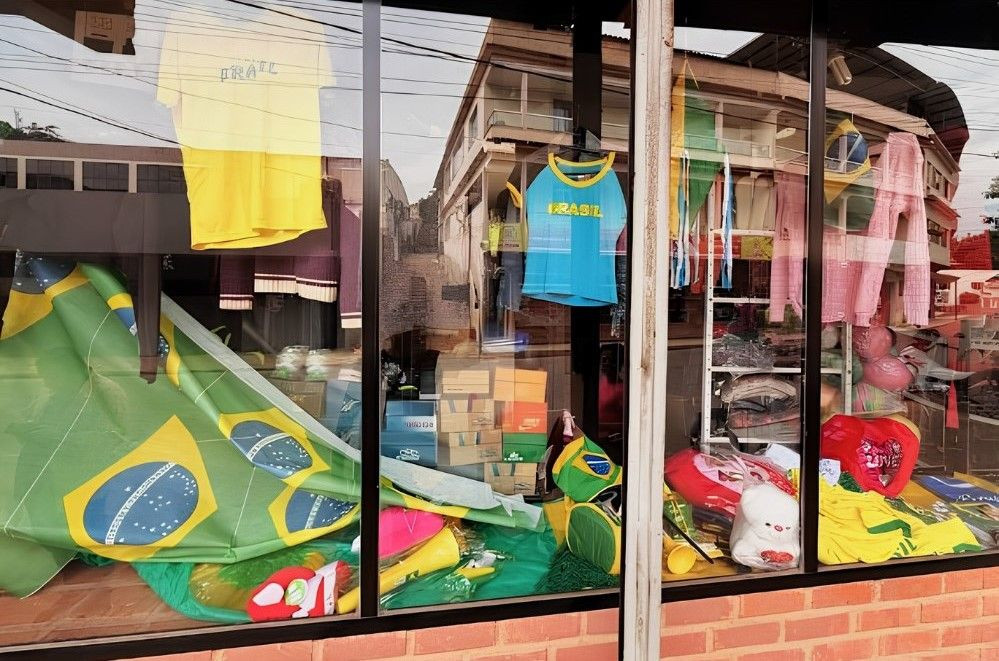 Reflexo na vidraça transparente de uma loja comercial no distrito. No interior da vitrine, estão expostas camisetas com as cores do Brasil (uma amarela e outra azul com detalhes amarelos), uma grande bandeira nacional estendida, cornetas de plástico amarelas e verdes, pelúcias e caixas de calçados organizadas ao fundo. O vidro reflete o casario e o relevo da rua oposta.