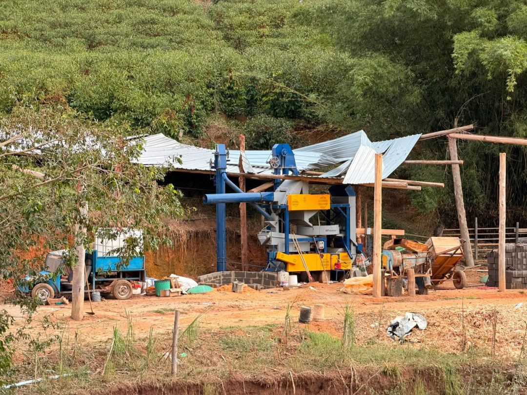 Fotografia em plano geral de uma propriedade rural mostrando uma máquina agrícola de grande porte, nas cores azul e amarela, posicionada sob uma estrutura de galpão que teve o telhado de zinco retorcido e parcialmente destruído pela tempestade. Ao fundo, uma encosta com vegetação verde e plantações de café.