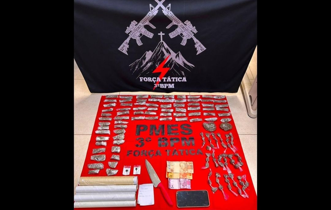 Foto de exibição de material apreendido pela polícia. Ao fundo, uma bandeira preta traz o emblema da 
