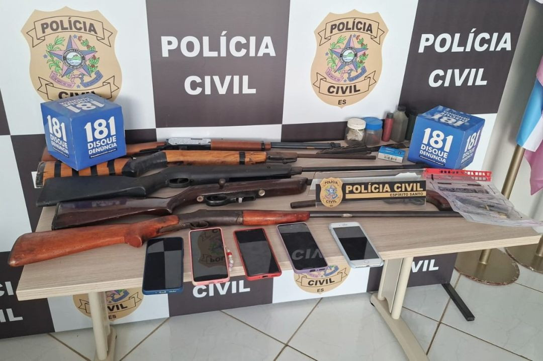 Fotografia horizontal mostrando o resultado de uma apreensão policial disposta sobre uma mesa de madeira clara. Ao fundo, há um painel xadrez em preto e branco com o brasão oficial e a inscrição 
