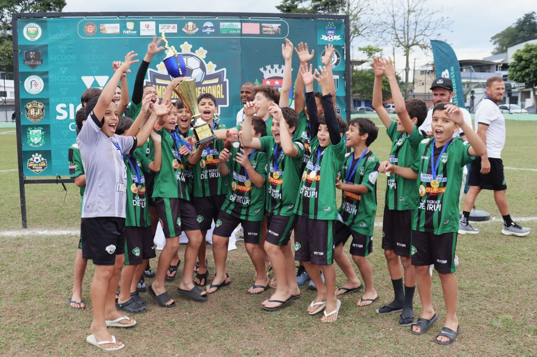Grupo de jogadores de futebol infantil da equipe de Irupi, vestindo uniforme verde e preto listrado com shorts pretos, comemorando a conquista do troféu. Os atletas sorriem, levantam os braços e exibem medalhas no peito em frente a um painel oficial do Campeonato Regional.