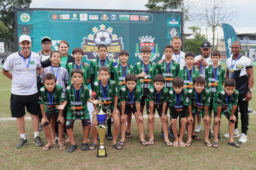 Foto oficial do time completo de Irupi posando enfileirado no campo. Os atletas da categoria de base usam camisas verdes com listras pretas e medalhas de ouro. À frente deles, o troféu de campeão está posicionado no gramado. Adultos da comissão técnica e organização acompanham o registro nas laterais e atrás.
