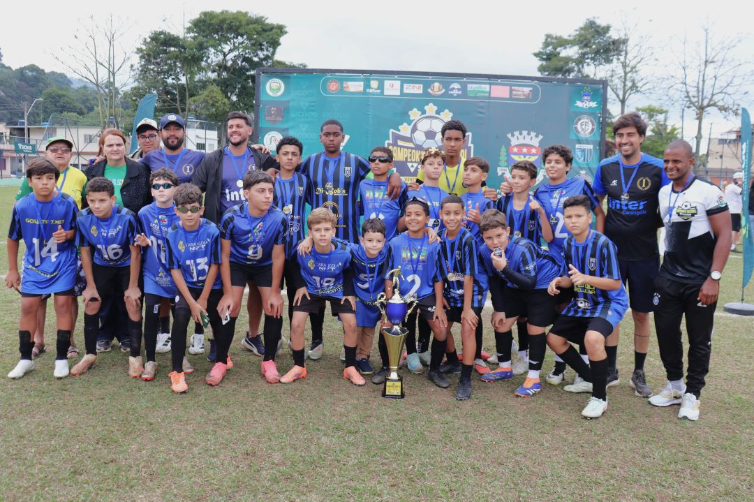 Jogadores da categoria Sub-13 da equipe Inter Carangola posam reunidos no gramado. Eles vestem uniforme azul com listras pretas verticais. À frente do grupo, centralizado no chão, está o troféu de campeão dourado e azul. Comissões técnicas e organizadores aparecem atrás dos atletas.