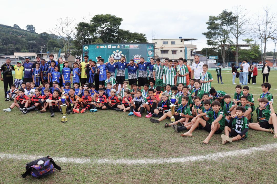 Foto panorâmica do gramado reunindo todas as equipes participantes, atletas juvenis e comissões técnicas após o encerramento do Campeonato Regional Infantil em Dores do Rio Preto. Jogadores vestindo diferentes uniformes (verde, azul, listrado vermelho e preto) aparecem sentados e em pé ao redor dos troféus dispostos no campo.