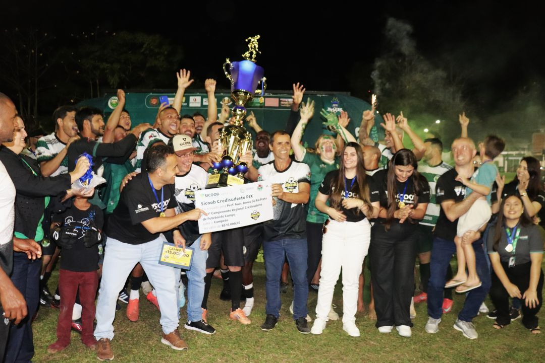 Grupo de jogadores e comissão técnica do time Jobalu comemorando no gramado à noite. Eles seguram um grande troféu azul e dourado e um cheque simbólico gigante do Sicoob Credisudeste no valor de R$ 15.000,00, destinado ao campeão. Há homens com camisas listradas em verde e branco e outros com camisas pretas da organização.