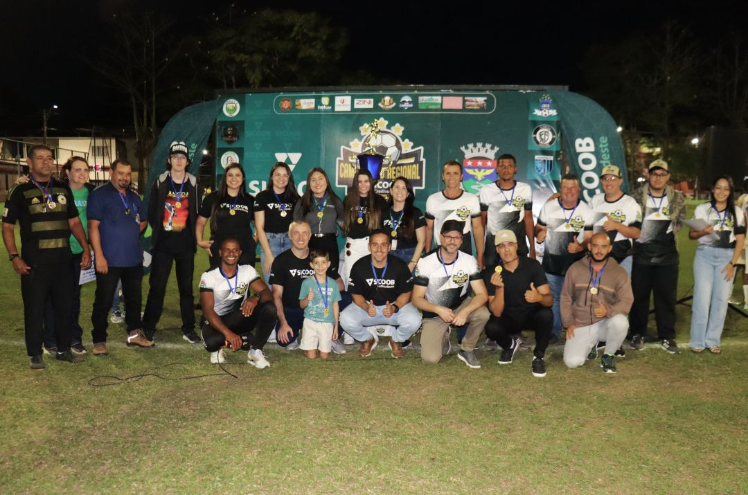 Registro noturno de organizadores, autoridades e representantes do Sicoob Credisudeste reunidos no campo após a premiação. Eles estão posicionados em frente ao portal inflável do evento, vestindo camisas pretas e brancas da organização e exibindo medalhas no peito. Uma criança pequena aparece no centro da imagem segurando uma medalha.