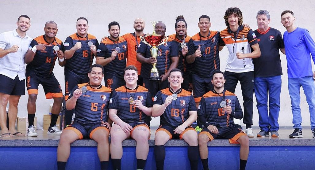 Foto grupal da equipe de handebol Tigers, de Rio Novo do Sul, comemorando no pódio do Ginásio de Esportes de Iúna. Os atletas vestem uniforme preto com listras cinzas horizontais e detalhes em laranja. No centro, um jogador segura um troféu grande, dourado e vermelho. Outros atletas sorriem segurando suas medalhas de ouro. À direita e à esquerda, integrantes da comissão técnica posam junto ao time.
