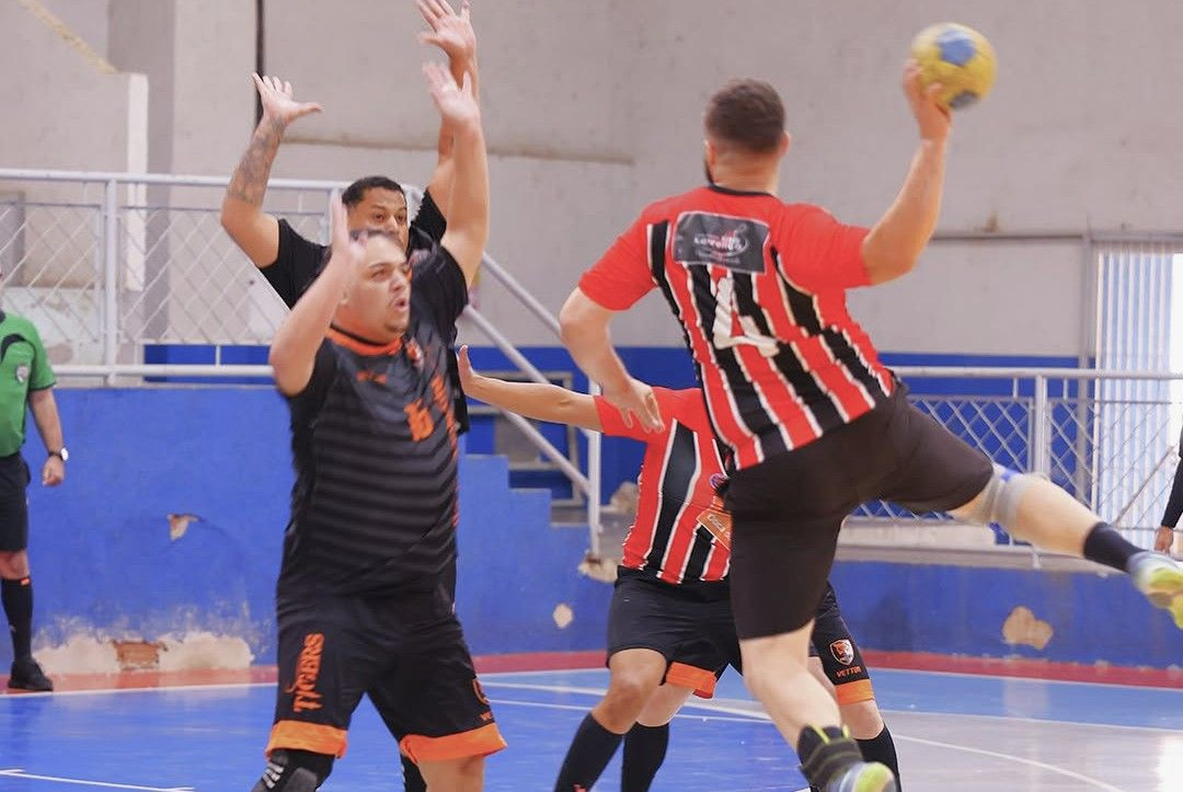 Registro de uma jogada de ataque durante a partida de handebol. Um jogador com uniforme listrado em vermelho e preto salta em suspensão, de costas para a câmera, preparando-se para arremessar a bola com a mão direita. Três defensores do time Tigers, vestidos de preto e laranja, estão posicionados com os braços levantados tentando bloquear o arremesso dentro da área de defesa azul.