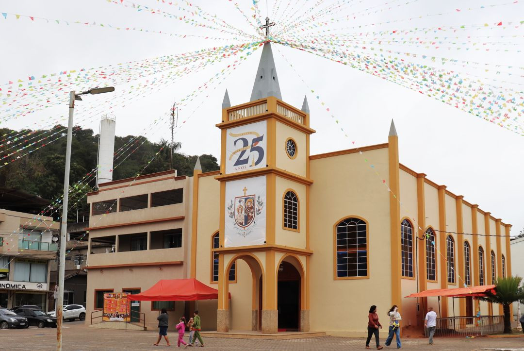 Fotografia da fachada da Igreja Matriz São João Batista em Irupi, sob céu nublado. A edificação possui arquitetura com paredes pintadas em tom amarelo-creme e detalhes estruturais em laranja-escuro. Na torre central, há um banner vertical branco com o logotipo comemorativo escrito 