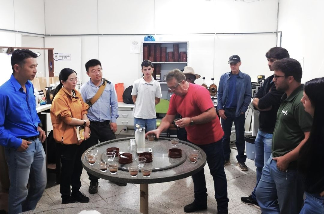 Grupo de pessoas reunido em uma sala técnica de classificação de café em Muniz Freire. No centro, ao redor de uma mesa redonda de granito com xícaras de café e amostras de grãos para degustação, um homem com camisa vermelha e óculos de proteção manipula uma das amostras. À esquerda, os pesquisadores chineses — um homem de camisa azul e uma mulher de casaco laranja — observam atentamente o procedimento de análise sensorial.