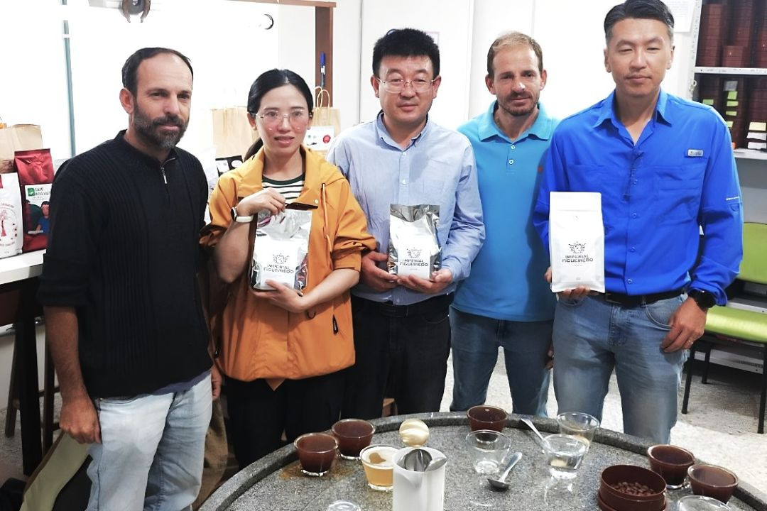 Cinco homens e uma mulher posam alinhados para foto atrás de uma mesa redonda com xícaras de café em processo de degustação. A pesquisadora chinesa, de casaco laranja, e os homens ao seu lado seguram pacotes de café especial da marca 