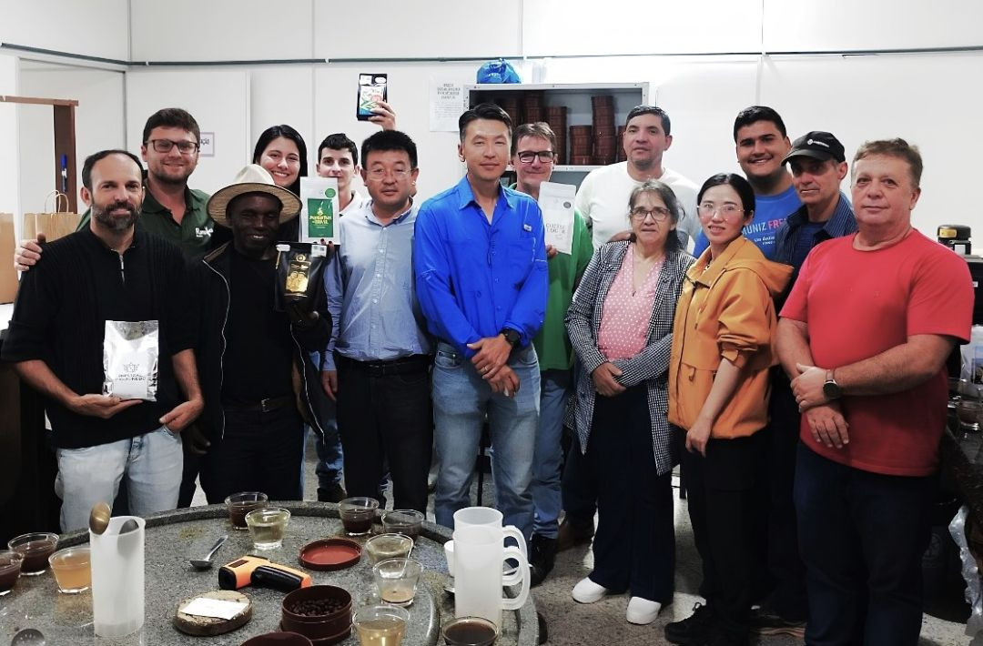 Foto oficial com o grupo completo de produtores, técnicos e autoridades locais reunidos junto aos pesquisadores chineses na Sala de Classificação de Cafés de Muniz Freire. Todos sorriem para a foto atrás da mesa de granito que contém utensílios de classificação, xícaras, bules plásticos e um termômetro digital infravermelho. Alguns produtores ao fundo seguram pacotes de marcas locais de café.
