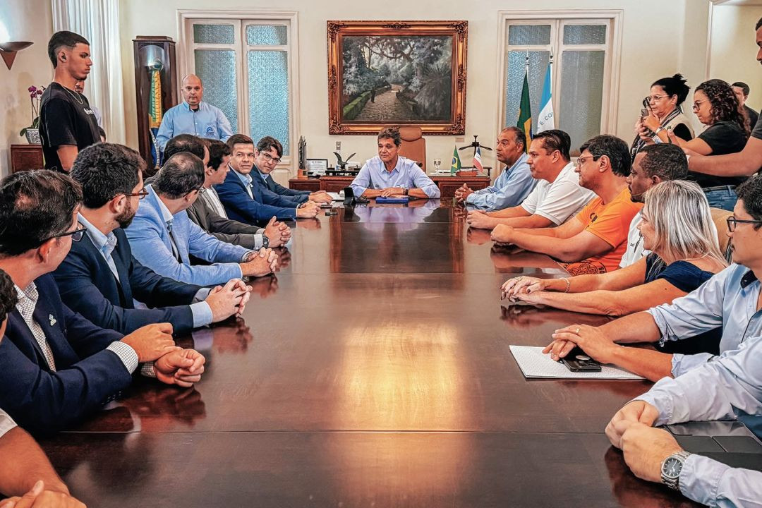 Fotografia em plano médio de uma reunião formal em uma sala com paredes claras e portas de vidro ao fundo. No centro da imagem, há uma grande mesa de madeira escura e polida que reflete a iluminação do ambiente. Cerca de vinte pessoas estão distribuídas ao redor da mesa. Ao centro, na cabeceira ao fundo, senta-se o governador do Espírito Santo, Ricardo Ferraço, vestindo camisa azul clara de mangas compridas dobradas. Nas laterais da mesa, homens e mulheres acompanham a reunião; alguns vestem camisas sociais e paletós, outros vestem camisetas casuais. No lado direito da imagem, duas mulheres em pé registram o momento com telefones celulares. No centro da parede ao fundo, há um quadro com moldura dourada retratando uma paisagem arborizada, flanqueado pelas bandeiras do Brasil e do Espírito Santo.