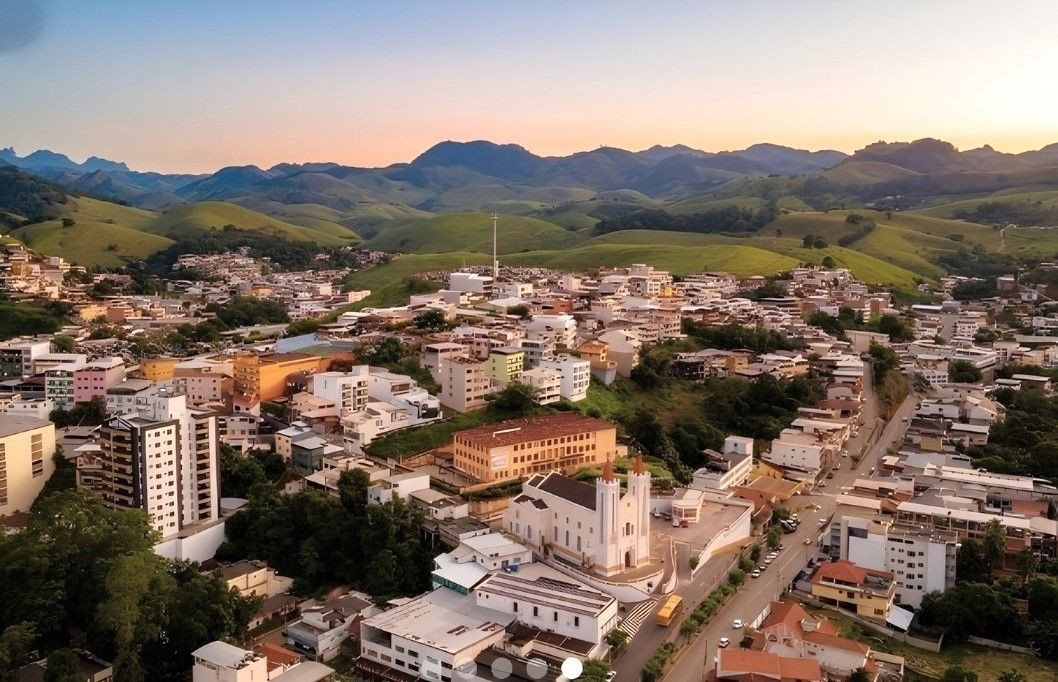 Vista aérea panorâmica da cidade de Alegre, no Espírito Santo, durante o final da tarde. Em primeiro plano, destacam-se o casario urbano, prédios residenciais e a Igreja Matriz de Nossa Senhora da Penha, com sua arquitetura branca e torres simétricas. Ao fundo, o relevo característico da região do Caparaó apresenta colinas verdes e montanhas sob um céu com tons suaves de laranja e azul claro.