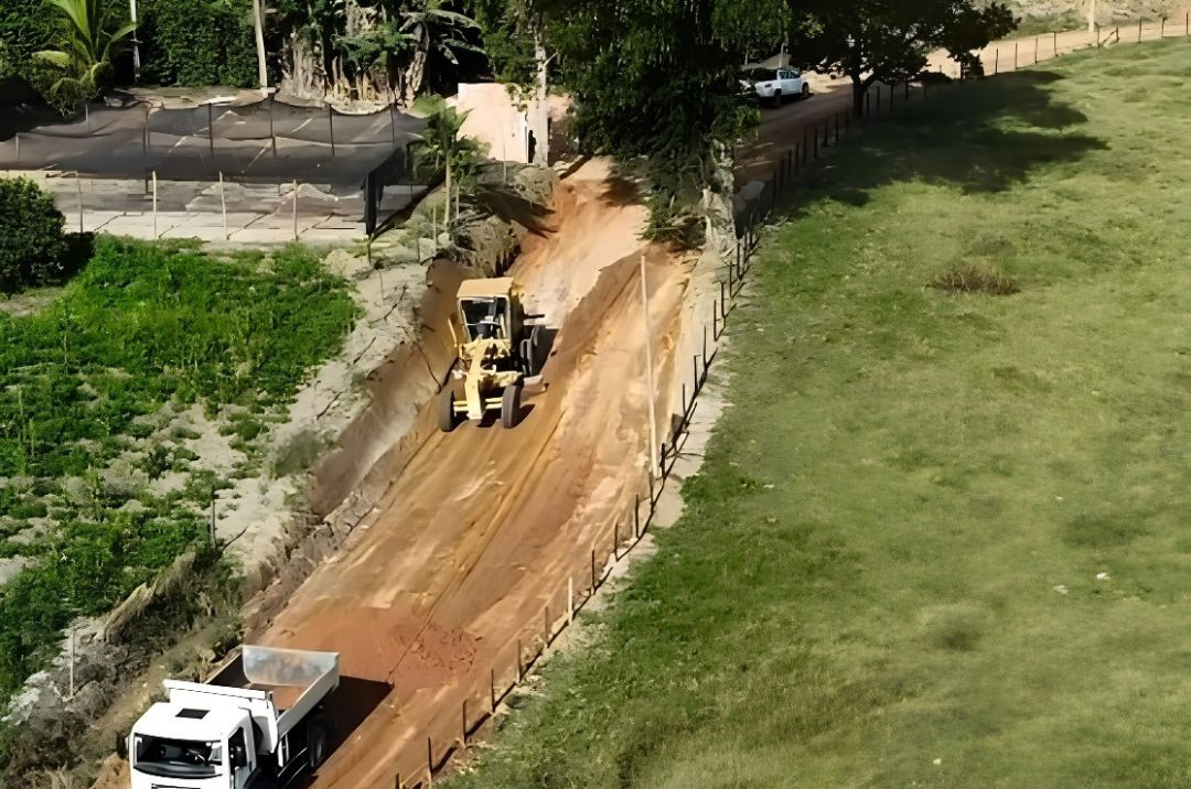 Vista aérea de uma estrada de terra em manutenção em Dores do Rio Preto. No centro, uma máquina motoniveladora (patrol) amarela trabalha no nivelamento da pista de solo avermelhado. Na parte inferior esquerda, a cabine de um caminhão caçamba branco é visível. A estrada é ladeada por uma cerca à direita, que delimita um pasto verde, e por árvores e uma área cultivada à esquerda. Ao fundo, um veículo utilitário branco está estacionado na via.