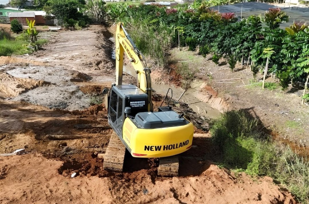 Fotografia em ângulo elevado mostrando uma escavadeira hidráulica amarela da marca New Holland operando na margem do Córrego Bom Jardim. A máquina está posicionada sobre a terra revirada, voltada para o leito do córrego onde realiza o desassoreamento. À direita, há vegetação rasteira e arbustos, enquanto ao fundo observam-se plantações e estruturas cobertas por telas sombreadoras pretas.
