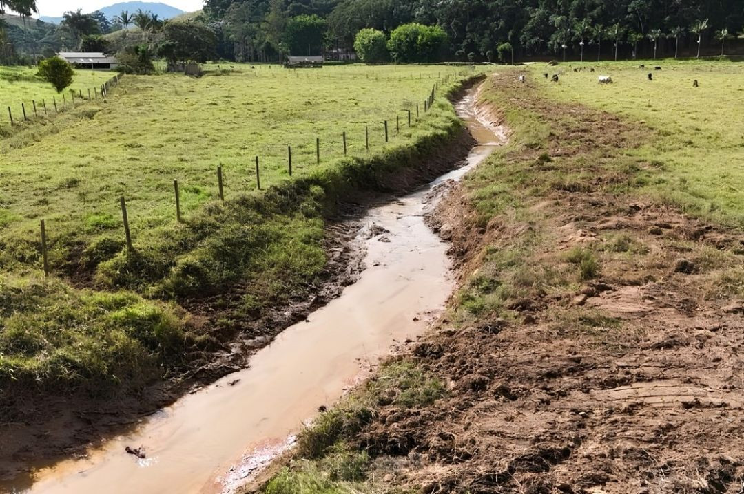 Imagem em perspectiva de um trecho sinuoso do Córrego Bom Jardim após a intervenção de limpeza. O leito de água barrenta corre entre margens de terra recentemente escavadas e limpas. À esquerda do córrego, há uma propriedade delimitada por estacas de madeira e cerca de arame, cobrindo uma vasta área de pastagem verde com árvores ao fundo. Gados pastam dispersos no campo à direita.