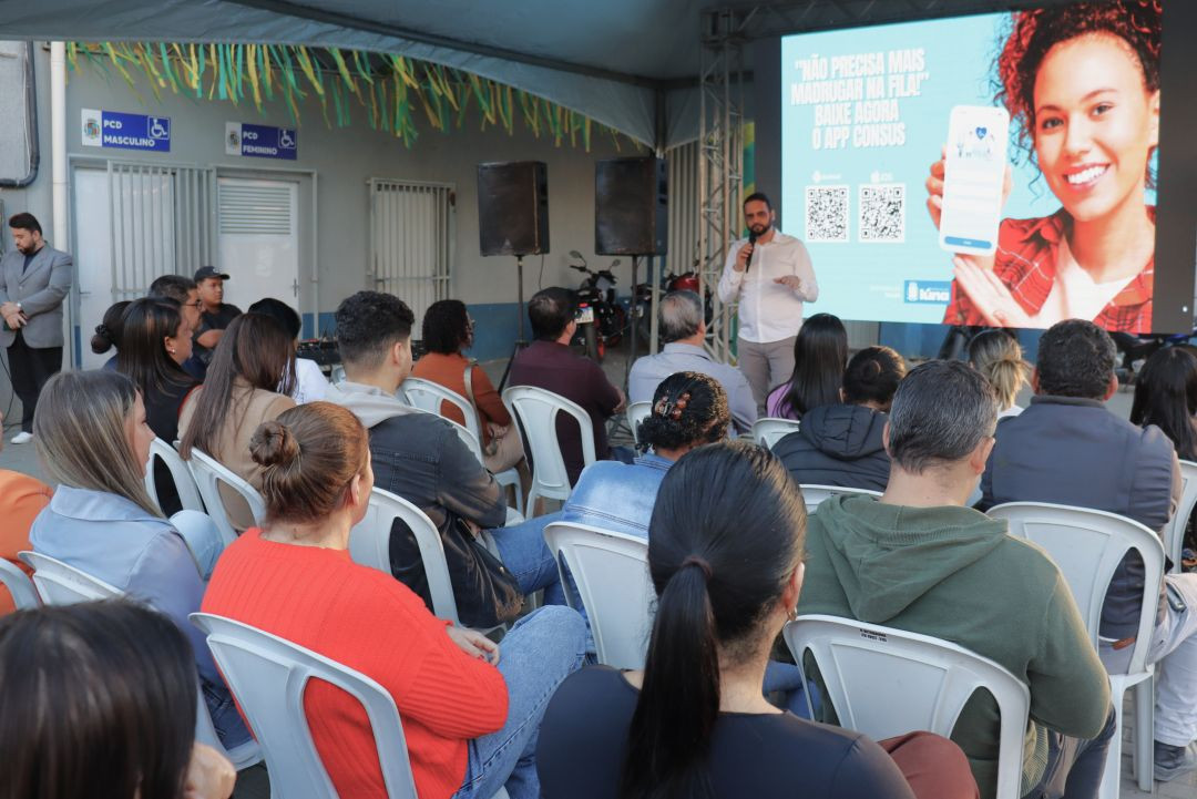 Vista da plateia sentada em cadeiras brancas sob uma tenda azul, assistindo à apresentação do aplicativo. Ao fundo, um homem de camisa branca fala ao microfone ao lado de uma grande tela digital que exibe a imagem de uma mulher sorrindo e segurando um celular com o aplicativo Consus, além de dois QR Codes. Ao lado esquerdo, placas indicam banheiros acessíveis (PCD Masculino e Feminino).