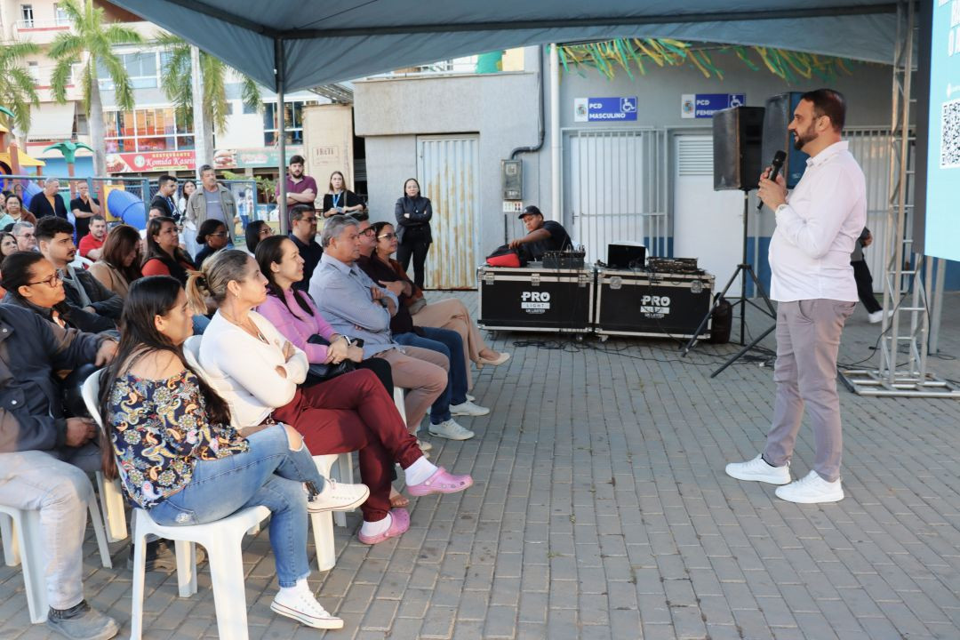 Homem de camisa social branca e calça cinza gesticula enquanto fala ao microfone para o público presente. Ele está posicionado de perfil em relação à câmera. A plateia, sentada em cadeiras brancas sob a estrutura da tenda, observa o pronunciamento. Ao fundo, é possível ver parte do comércio local e do Ginásio de Esportes de Iúna.