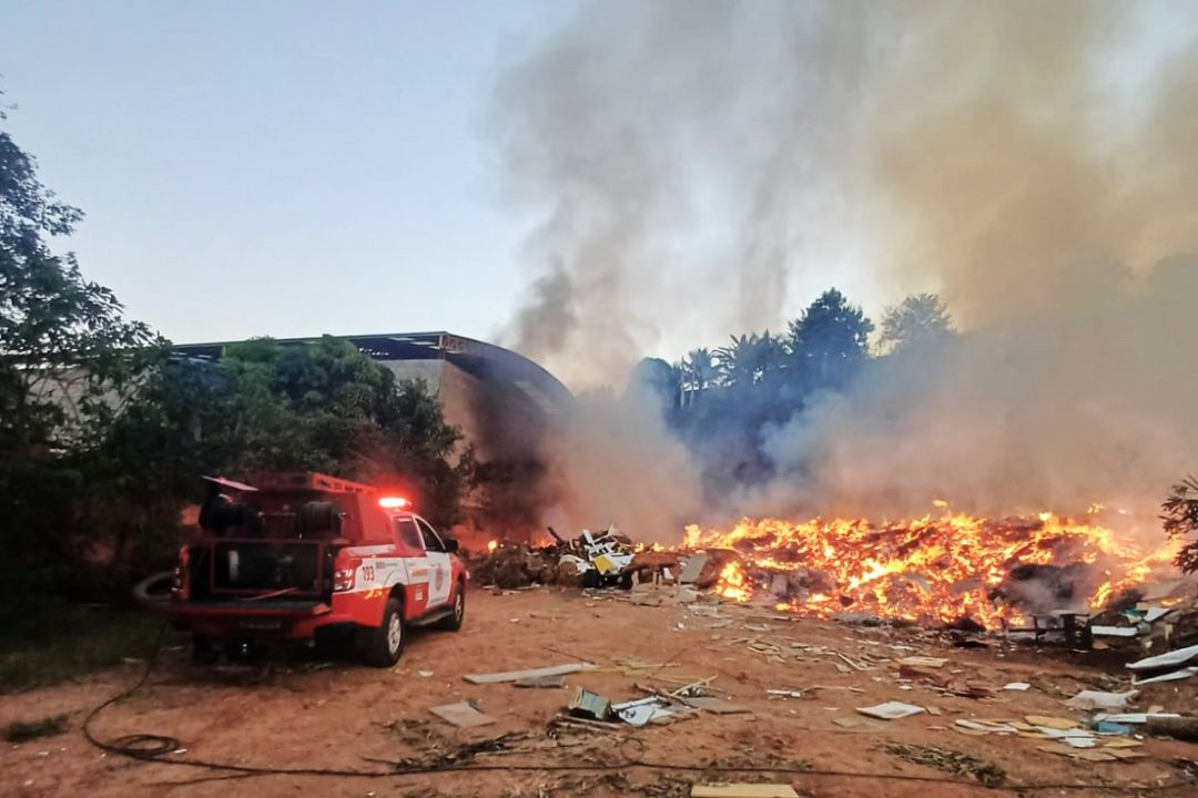 Viatura vermelha do Corpo de Bombeiros com as luzes de emergência ligadas estacionada em terreno de terra. Ao fundo, fumaça escura e focos de fogo consomem entulhos acumulados perto da estrutura de um galpão.