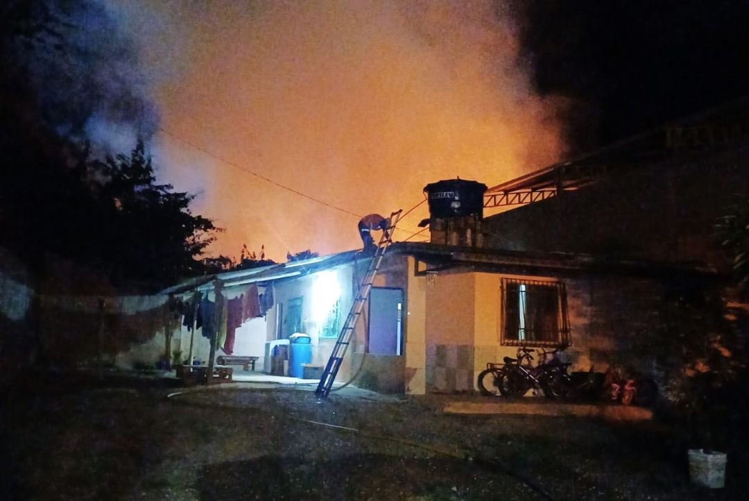 Visão noturna de um bombeiro ou socorrista no topo de uma escada apoiada no telhado de uma residência térrea iluminada. Ao fundo, o céu está avermelhado pelo reflexo do incêndio e coberto por fumaça.