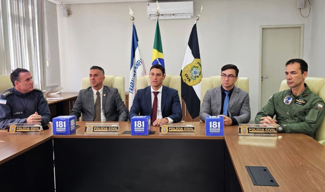 Cinco autoridades de segurança pública do Espírito Santo estão sentadas lado a lado atrás de uma mesa de madeira escura em uma sala de coletiva de imprensa. Ao fundo, estão posicionadas três bandeiras oficiais em pedestais: a do Estado do Espírito Santo, a do Brasil e a da Polícia Civil. Sobre a mesa, há placas de identificação escritas 