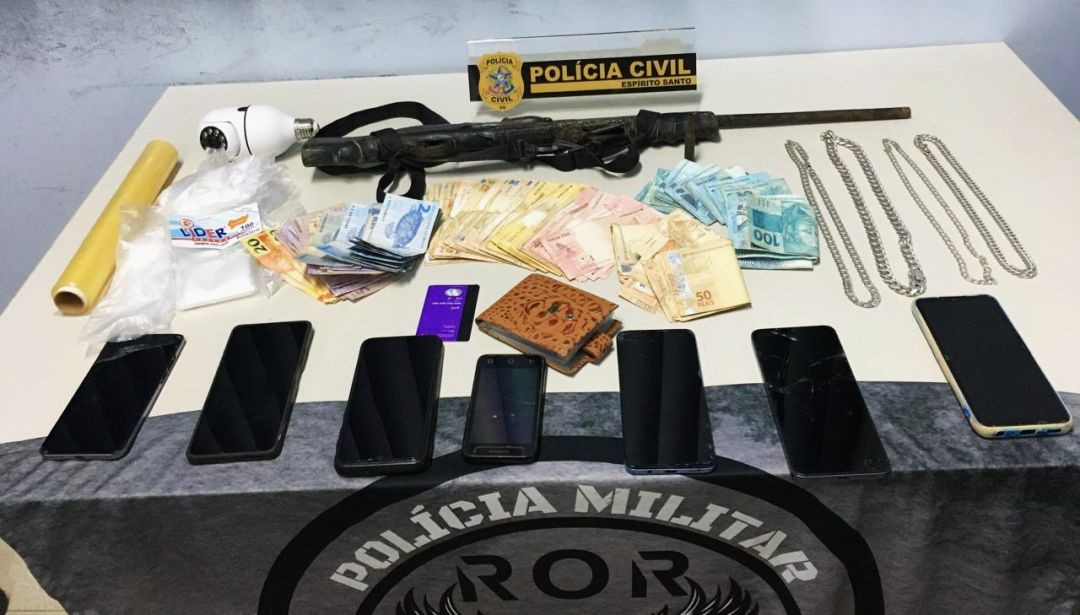 Sobre uma mesa clara coberta parcialmente por um tecido cinza com o brasão da Polícia Militar e a inscrição 