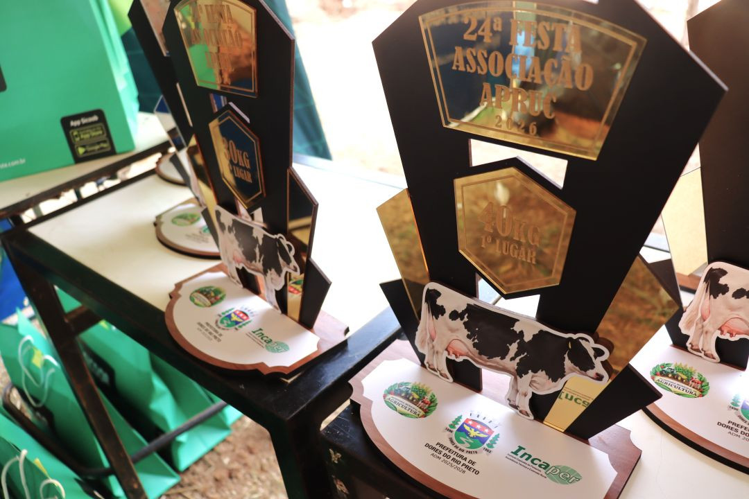 Fotografia em close-up focando em dois troféus de premiação dispostos sobre uma mesa na Festa do Cerro. Os troféus possuem design moderno em acrílico preto e dourado, cada um apresentando uma imagem recortada de uma vaca holandesa malhada. Placas douradas gravadas no topo de ambos exibem o texto 