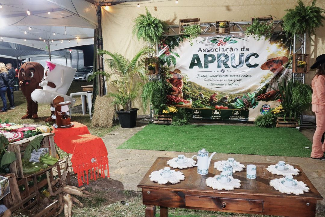 Fotografia de um espaço decorado em ambiente coberto na Festa do Cerro. Ao fundo, há um painel lona da Associação da APRUC com a imagem de lavouras de café e um produtor rural, emoldurado por estruturas metálicas com plantas samambaias e caixotes de madeira. À esquerda, dois bonecos de mascote fantasiados (um grão de café e uma xícara) interagem no local. Em primeiro plano, há uma mesa rústica de madeira comprida com bules, xícaras e pires de porcelana branca com detalhes azuis dispostos sobre toalhas de crochê brancas.