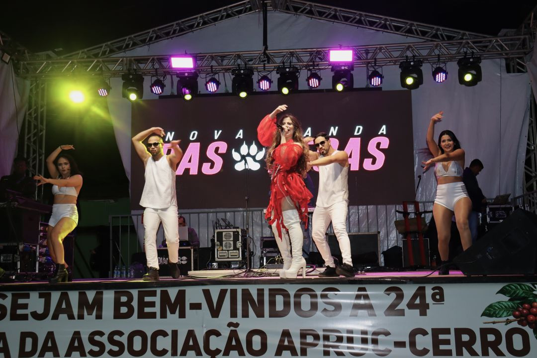 Fotografia de uma apresentação musical noturna no palco da Festa do Cerro. Uma cantora central, vestindo um vestido vermelho curto com franjas e botas brancas de cano alto acima do joelho, canta ao microfone. Ela é acompanhada por quatro dançarinos (dois homens e duas mulheres) vestidos com roupas brancas e óculos escuros, realizando uma coreografia. Ao fundo, um painel de LED exibe o nome da atração 