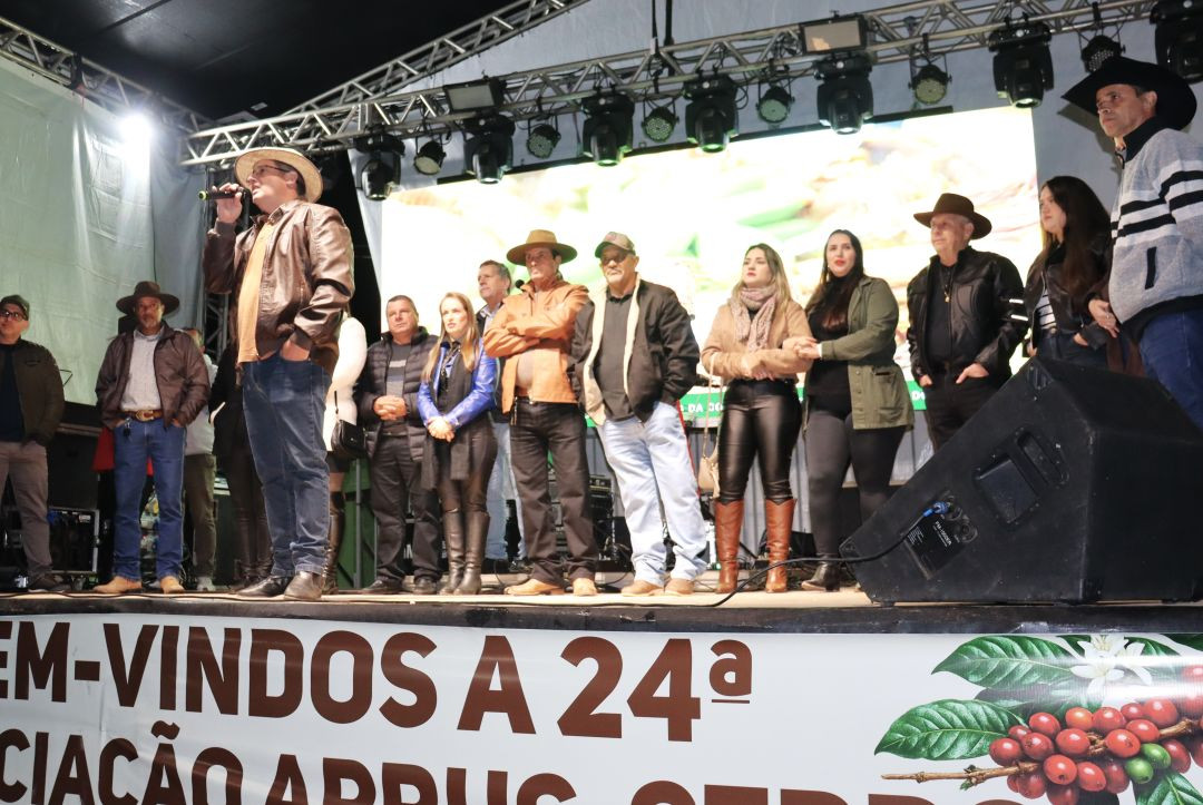 Fotografia do palco principal da festa sob iluminação noturna. Um grupo de cerca de doze pessoas, incluindo lideranças comunitárias e autoridades políticas vestindo casacos e chapéus de estilo sertanejo/rural, está alinhado lado a lado. À esquerda, um homem com chapéu de palha e jaqueta de couro marrom fala ao microfone. Na base do palco, uma lona branca traz os dizeres parciais 