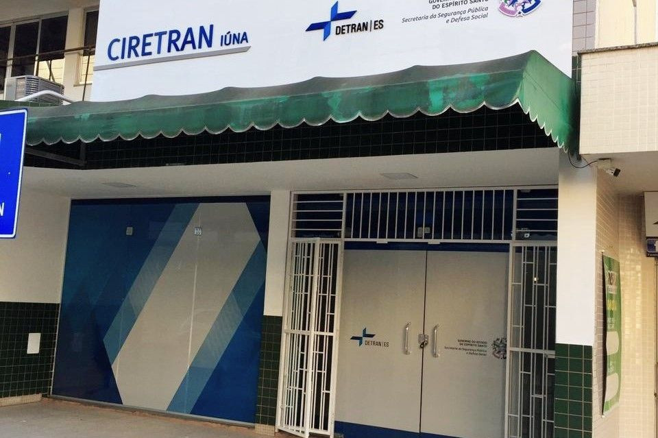 Fachada da Ciretran de Iúna, no Espírito Santo. O prédio possui uma cobertura verde estilo toldo na entrada, paredes brancas com detalhes em azulejos verdes escuros e portas de metal cinza fechadas. Na parte superior, há uma placa com a inscrição 