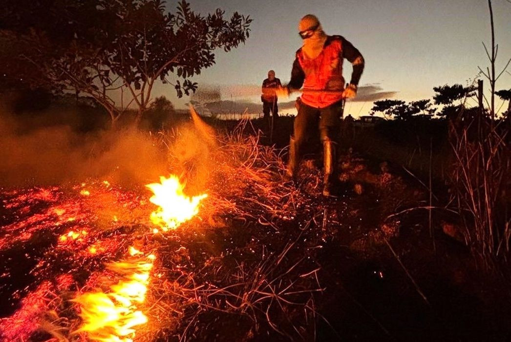 Fotografia em plano médio e ângulo normal, sob a luz do crepúsculo. Em primeiro plano, no canto inferior esquerdo, linhas de fogo consomem a vegetação seca e rasteira, gerando fumaça. Um brigadista, homem de pele clara vestindo calça escura, colete laranja reflexivo, luvas protetoras e balaclava clara cobrindo o rosto com óculos de proteção, caminha sobre a terra batida e usa uma ferramenta longa (abafador) para conter as chamas. Ao fundo, a silhueta de uma segunda pessoa observa a ação entre árvores e arbustos contra o céu escurecido com tons de laranja no horizonte.