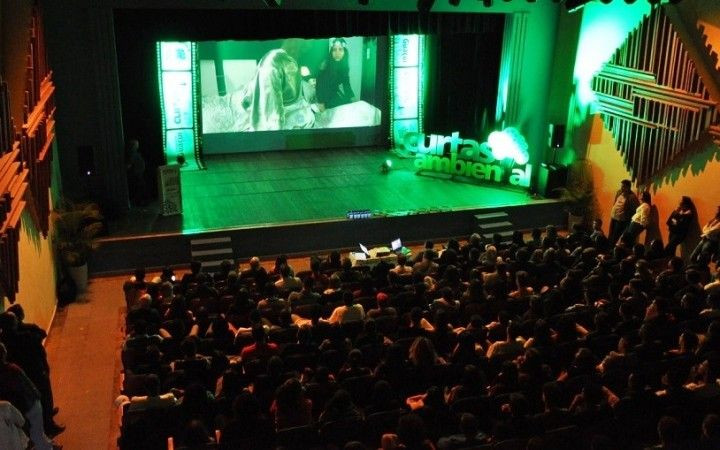 Imagem vertical colorida mostrando o interior do Teatro Municipal Fernando Torres em Guaçuí, ES. A plateia está repleta de pessoas, sentadas em poltronas escuras, voltadas para o palco. No palco, que possui iluminação verde predominante, há um telão grande onde é projetada uma cena de um vídeo (provavelmente um dos curtas). À direita do telão, no palco, está um banner vertical com a inscrição “CURTAS AMBIENTAL” e alguns logos, também com iluminação verde. À esquerda do telão, outro banner menor. O teto e as laterais do teatro têm detalhes arquitetônicos e técnicos, como luminárias e estruturas de metal. A foto captura a atmosfera do evento.