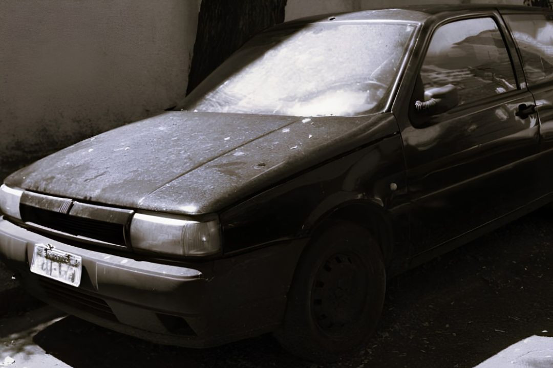 Foto em ângulo diagonal de um automóvel sedan preto com sinais de abandono, como sujeira e falta de conservação, estacionado em local que parece ser uma rua ou área de depósito, próximo a uma parede e um tronco de árvore.