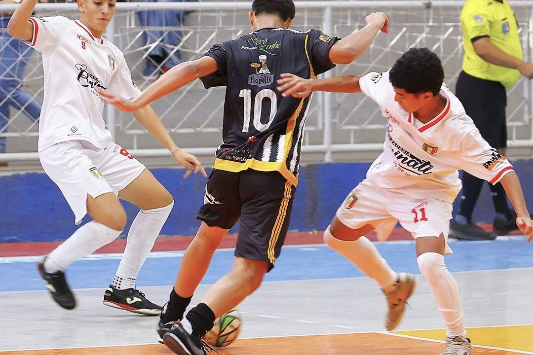 Foto vertical em ângulo fechado mostra uma disputa de bola em um jogo de futsal. Um jogador de costas, vestindo uniforme preto com listras amarelas e o número 10, protege a bola com os pés. Ele está cercado por dois adversários com uniforme branco e detalhes em vermelho (números 9 e 11) que tentam fazer a marcação. Ao fundo, uma grade de proteção e parte de um árbitro com camisa amarela.