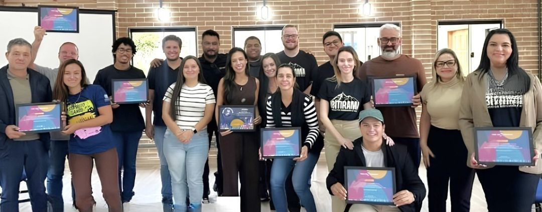 Foto em plano médio, horizontal, mostrando um grupo de aproximadamente 17 pessoas alinhadas em pé, sorrindo e posando para a foto em um espaço interno com parede de tijolos aparentes. Várias pessoas seguram certificados emoldurados em quadros pretos. O grupo veste roupas casuais, e uma mulher à direita veste um blazer bege.